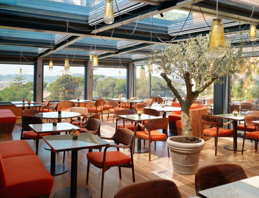 Glass-enclosed restaurant with vibrant orange chairs and tables, offering a beautiful view, creating a warm and inviting atmosphere.